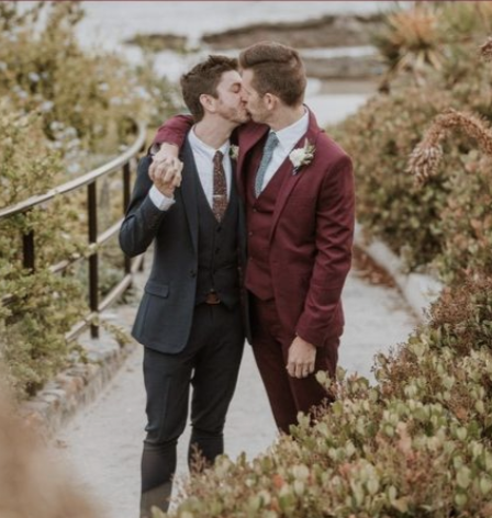 Two grooms in burgundy/dark suits kissing outdoors